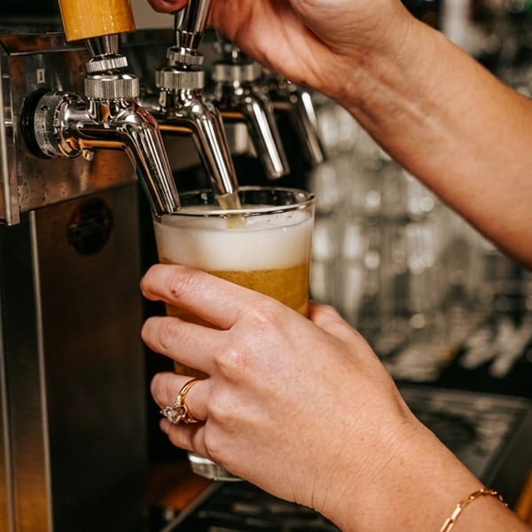 Hands pouring a golden beer with a frothy head from a tap into a clear glass.