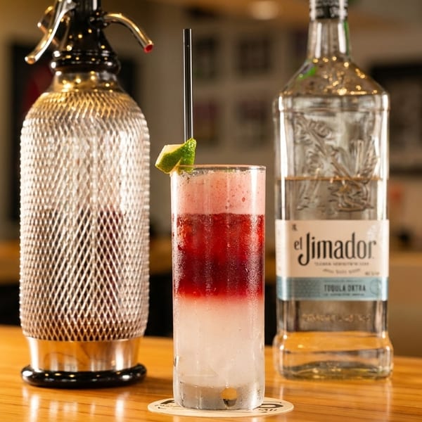 A layered red and white cocktail with a lime wedge, next to a tequila bottle and soda siphon.