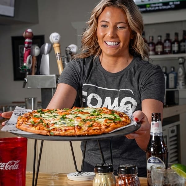 A smiling server in a grey FOMO shirt presents a fresh pizza on a stand at a bar.